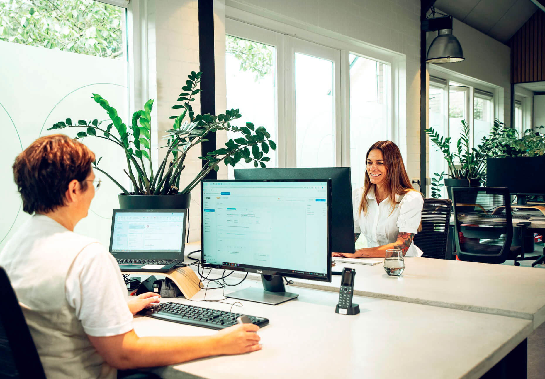2 collega's die aan het werk zijn achter hun laptop en ze glimlachen.